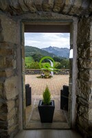 Un marco de piedra enmarca una puerta que da a un patio con una planta en maceta, muebles de mimbre y vistas a un paisaje montañoso.