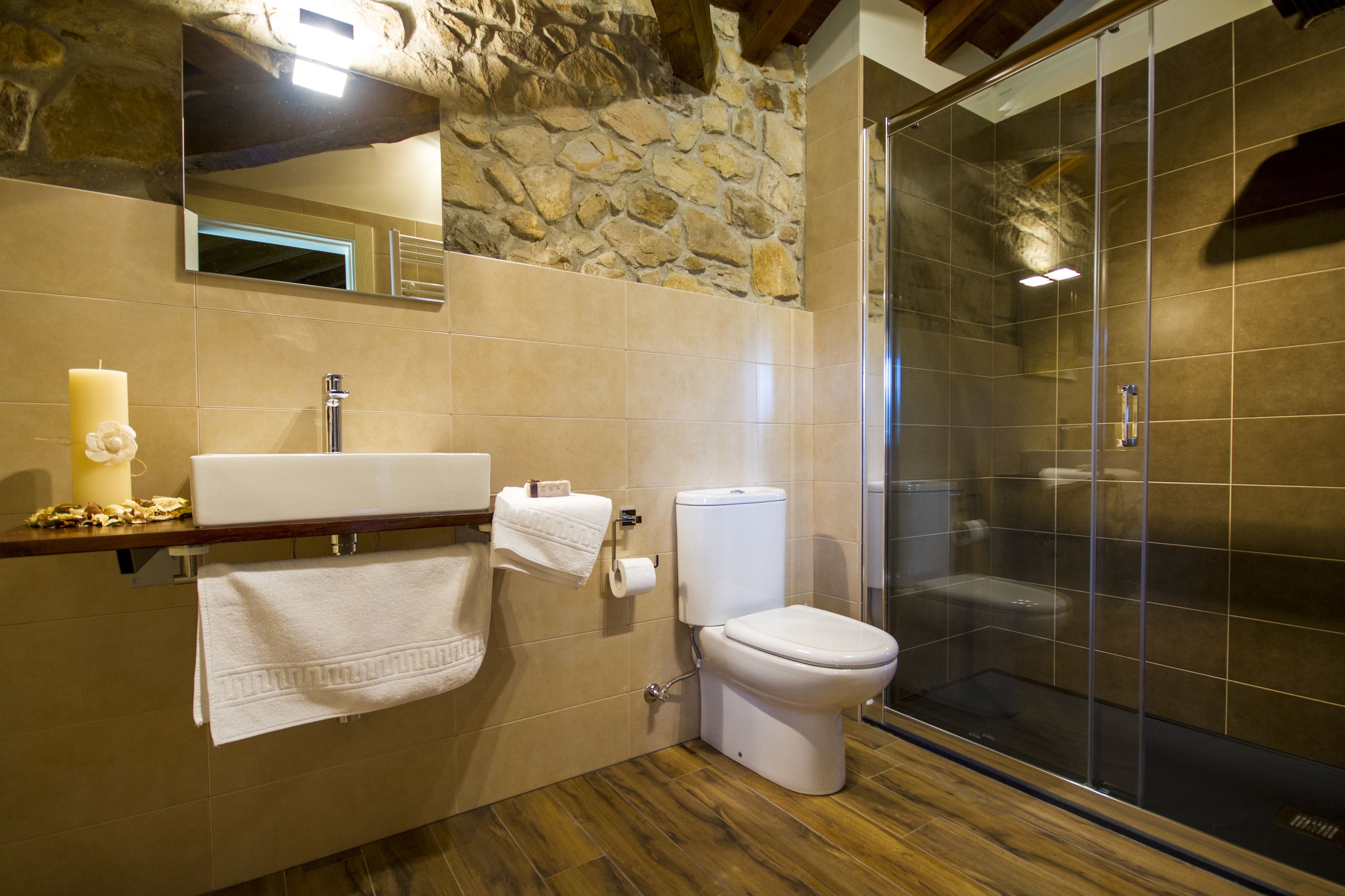 Une salle de bain avec un mur en pierre, un plancher en bois, un lavabo, des toilettes et une douche en verre.