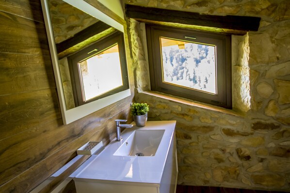 Un baño con pared de piedra, suelo de madera, lavamanos blanco y ventana con vista al cielo.