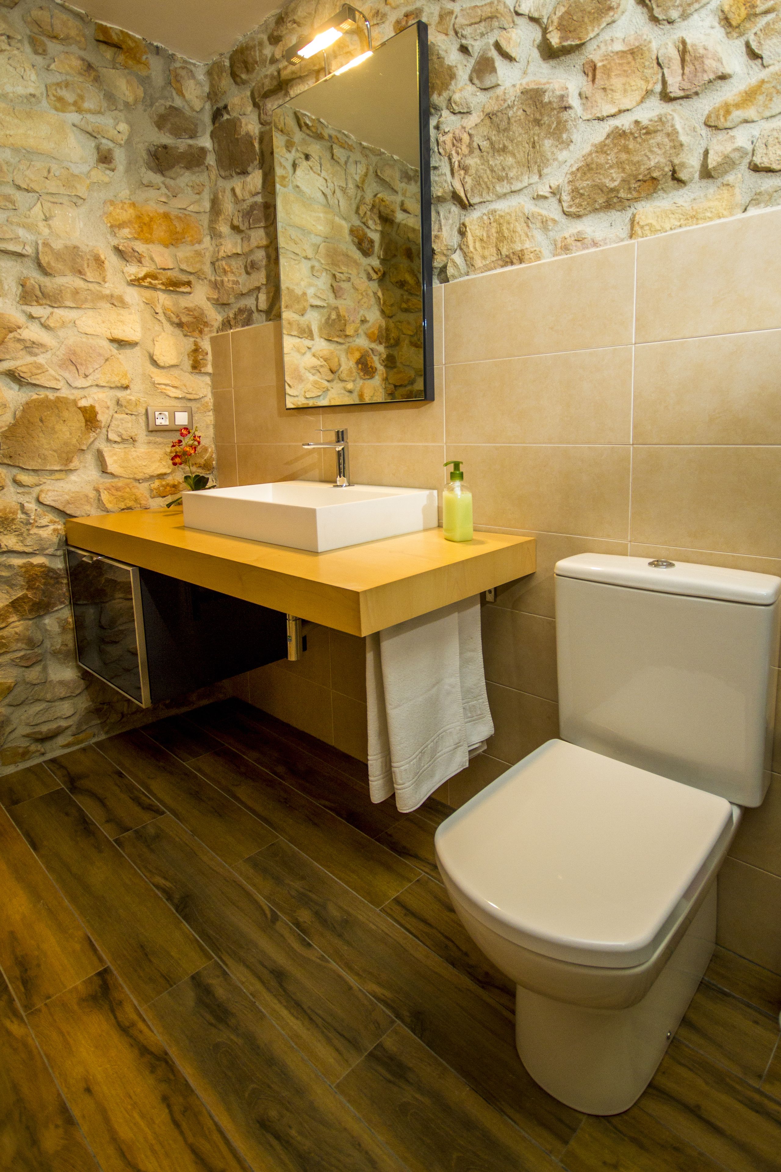 Un baño con pared de piedra, suelo de madera, lavamanos blanco, inodoro y espejo.