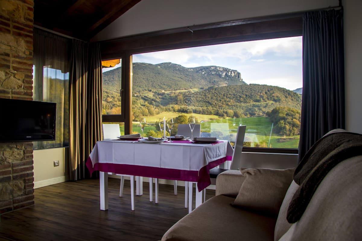 Un comedor con una mesa dispuesta para dos, un televisor instalado en una pared de piedra y vistas a un paisaje montañoso a través de grandes ventanales.