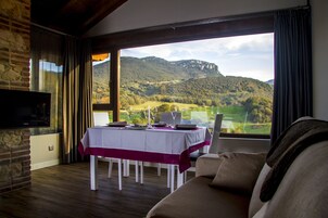 Un comedor con una mesa dispuesta para dos, un televisor instalado en una pared de piedra y vistas a un paisaje montañoso a través de grandes ventanales.