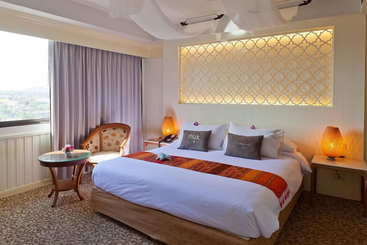 A hotel room with a large bed, a chair, a small table, a nightstand, and a decorative wall panel.