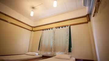 A room with two single beds, a wooden floor, a window with a floral curtain, and a ceiling light fixture.