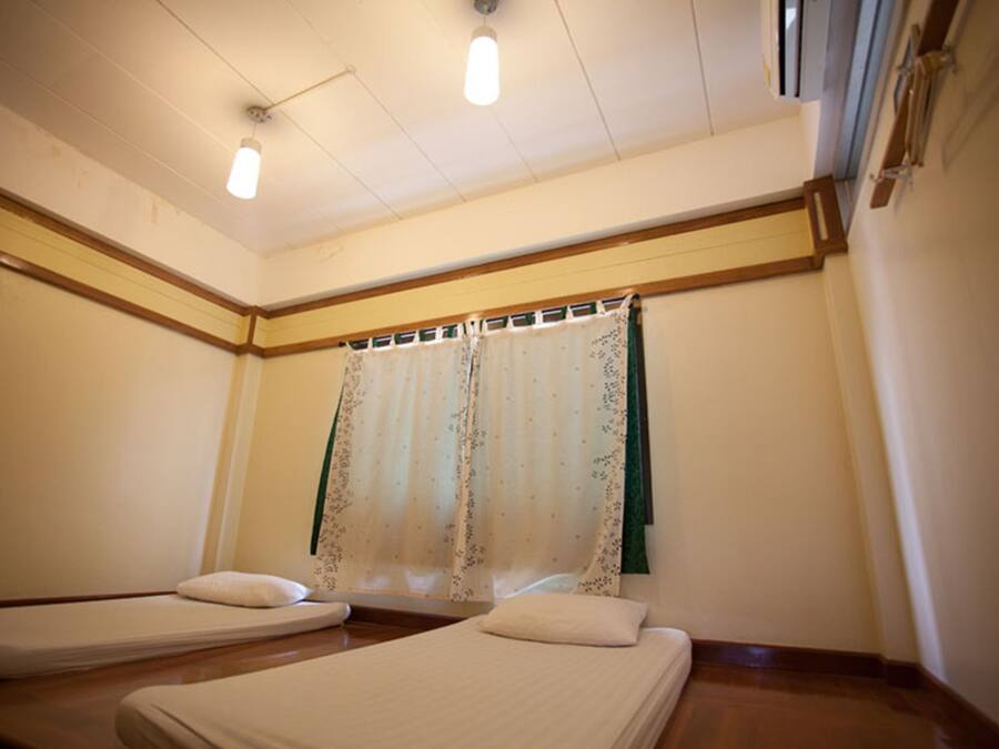 A room with two single beds, a wooden floor, a window with a floral curtain, and a ceiling light fixture.