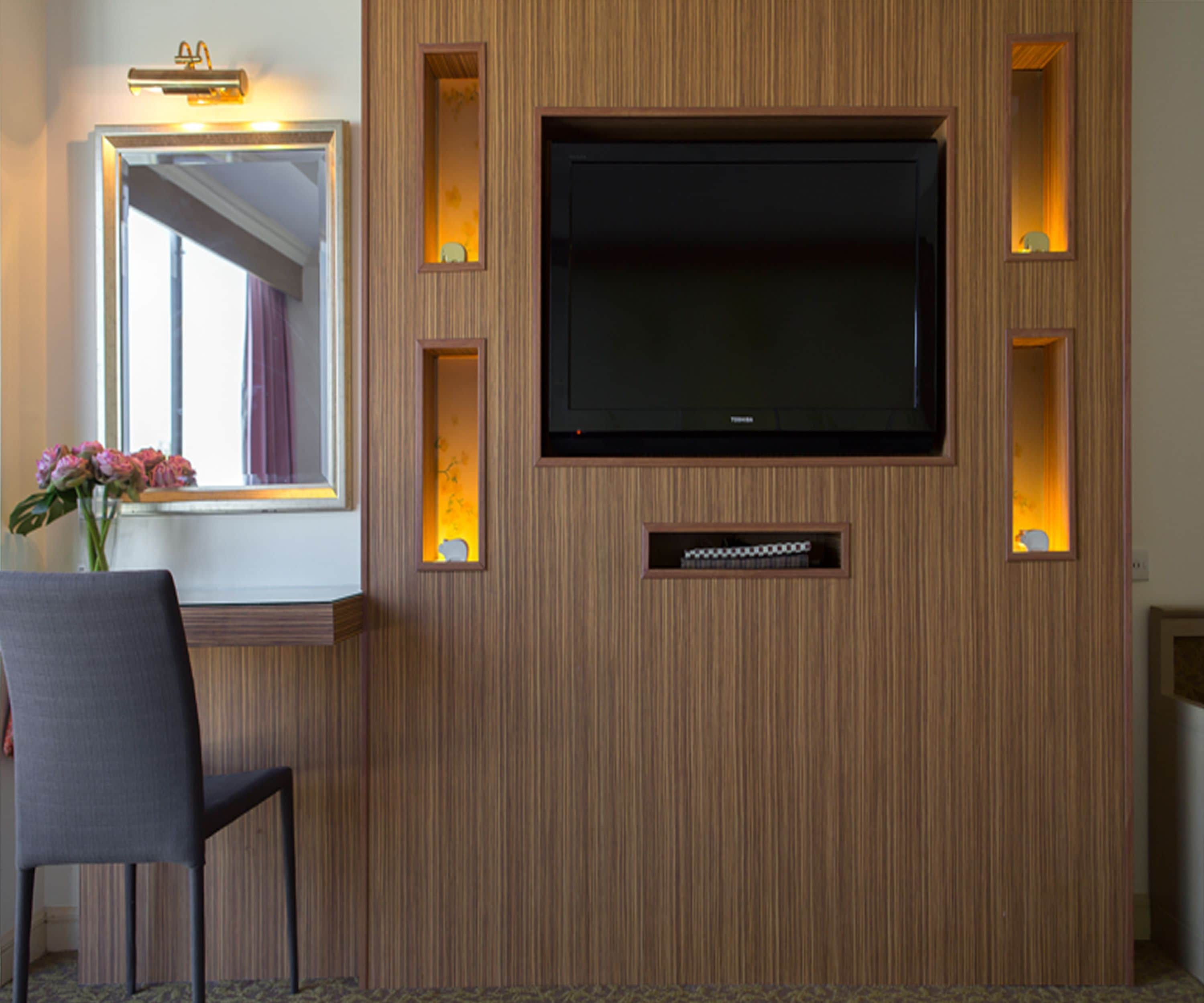 A hotel room with a mounted flat-screen TV, a mirror, and a chair.