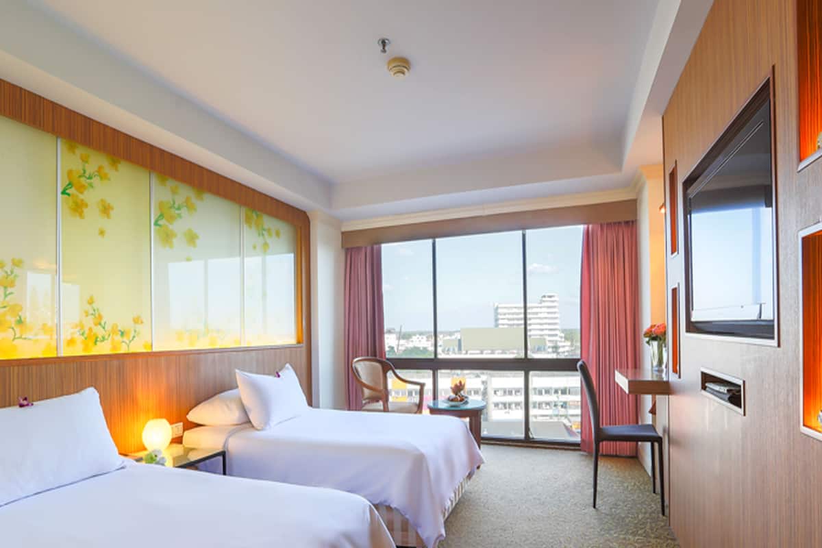 A hotel room with two beds, a desk, a chair, a TV, and a large window with a city view.