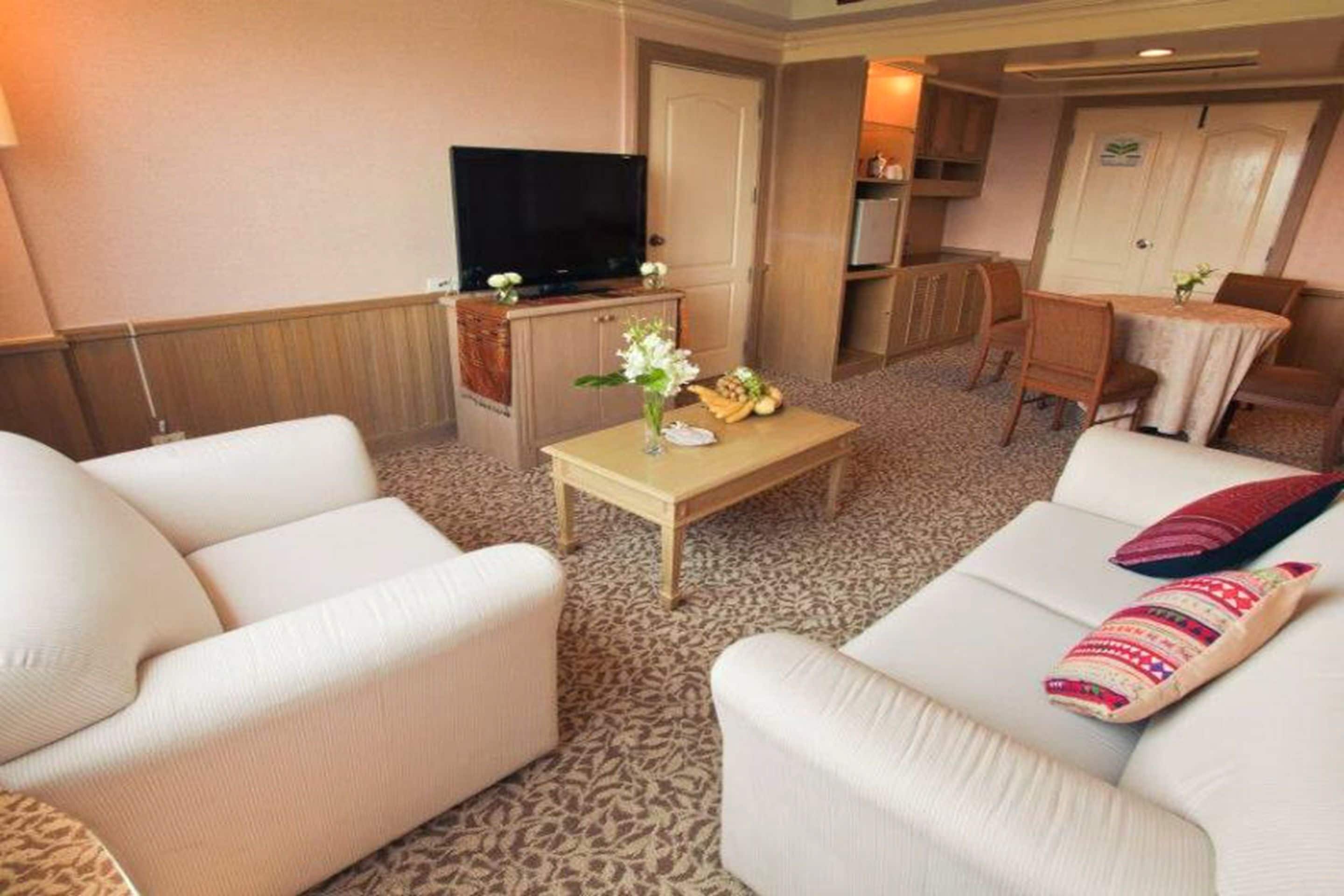 A hotel room with a sofa, armchairs, a TV, a small table with a vase, and a dining area with chairs and a table.
