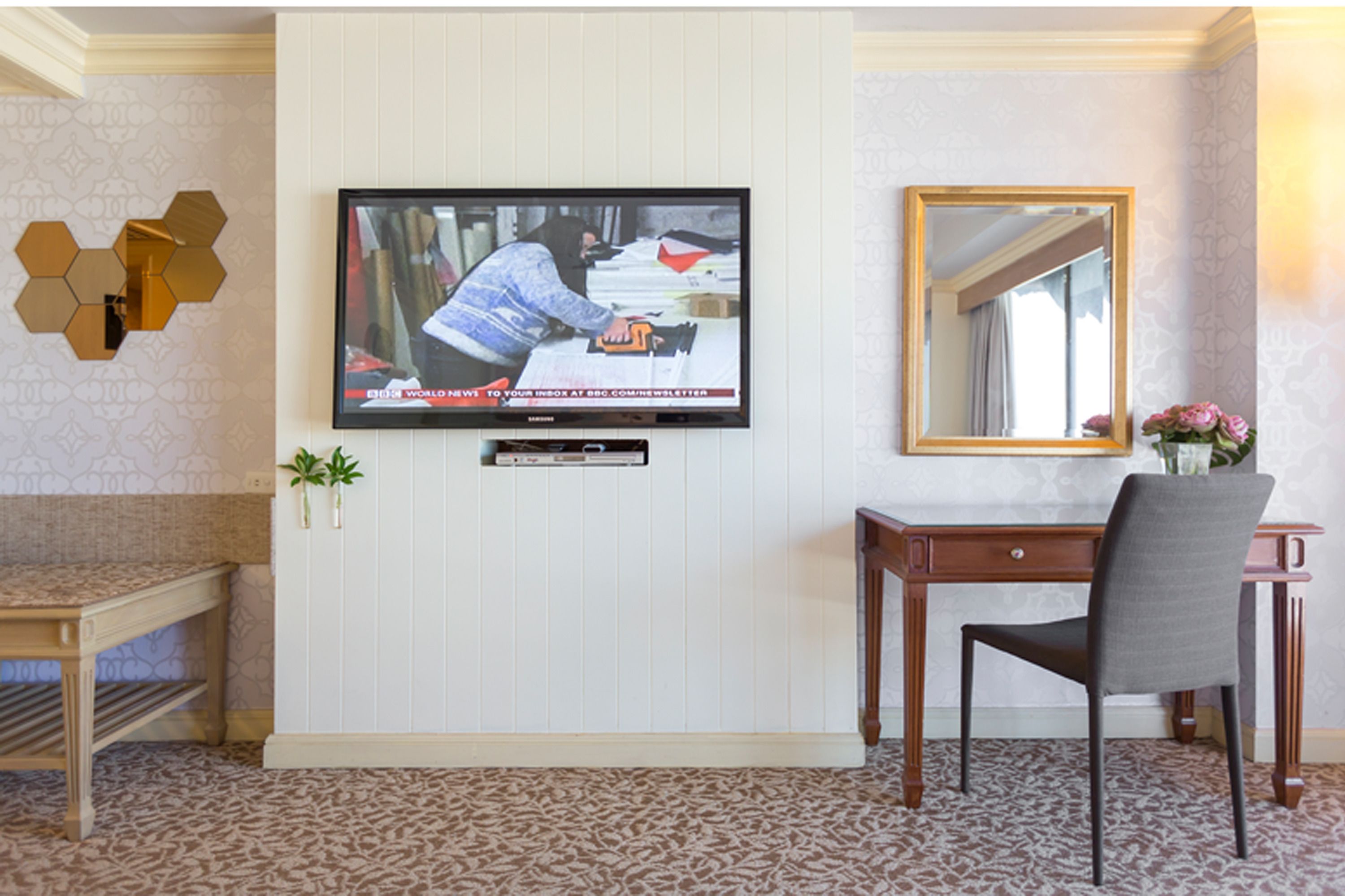 A room with a television mounted on the wall, a wooden desk, a chair, and a mirror.