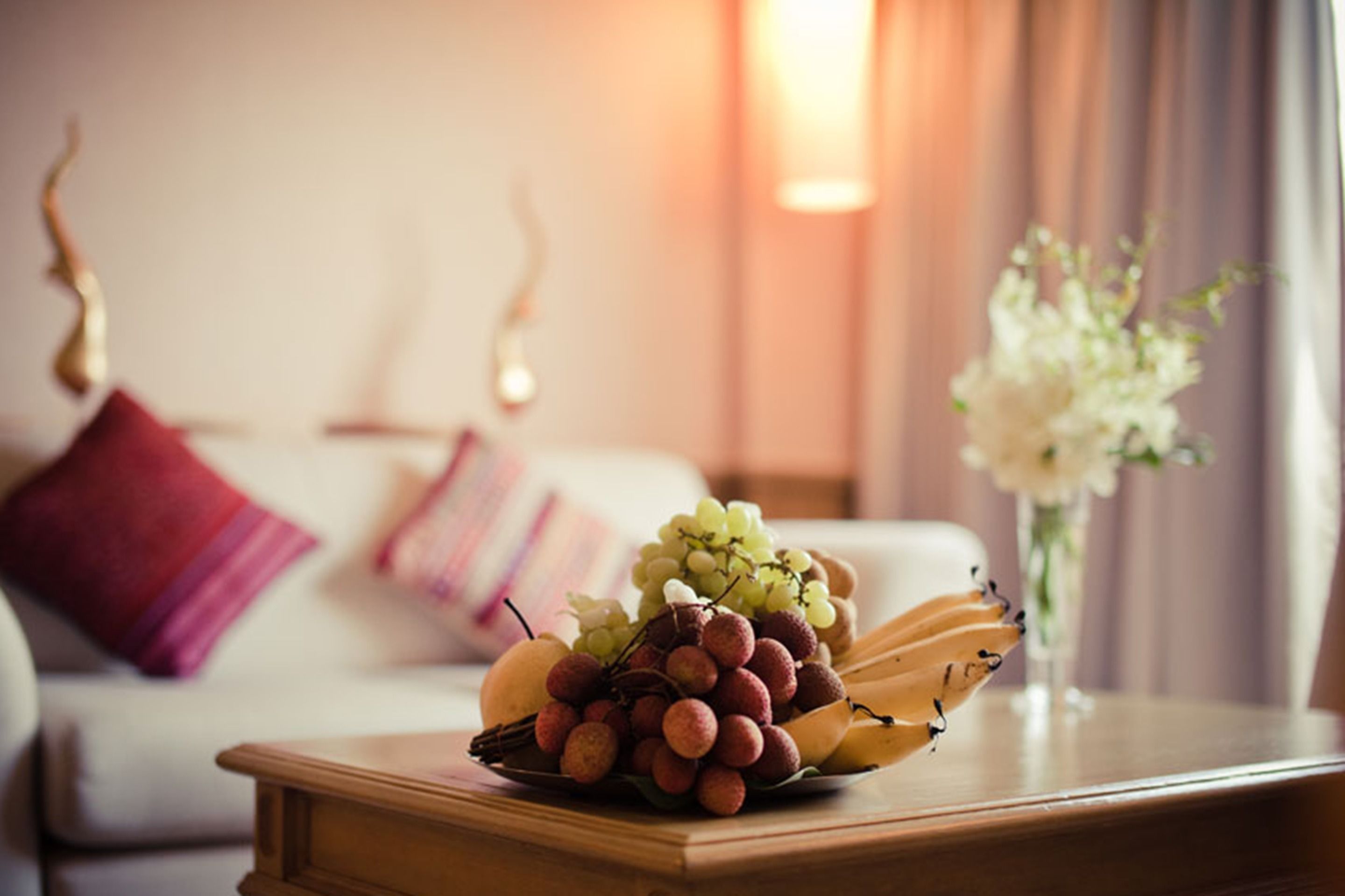 A fruit bowl on a wooden table with a bed and pillows in the background.