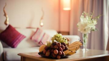 A fruit bowl on a wooden table with a bed and pillows in the background.
