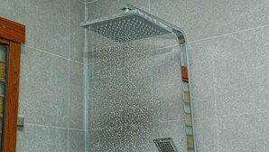 Une douche dotée d’un pommeau de douche imitant la pluie, d’une grille métallique et de murs recouverts de carreaux gris.