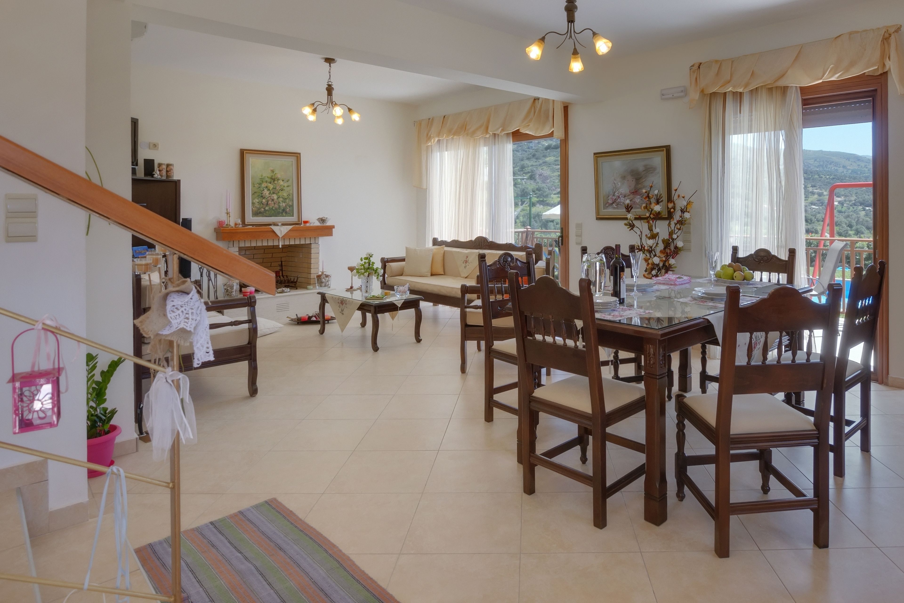 Un vaste espace de vie comprenant une table à manger, des chaises et un canapé. Un escalier se trouve sur la gauche, ainsi qu’une cheminée et un balcon offrant une vue sur les collines.