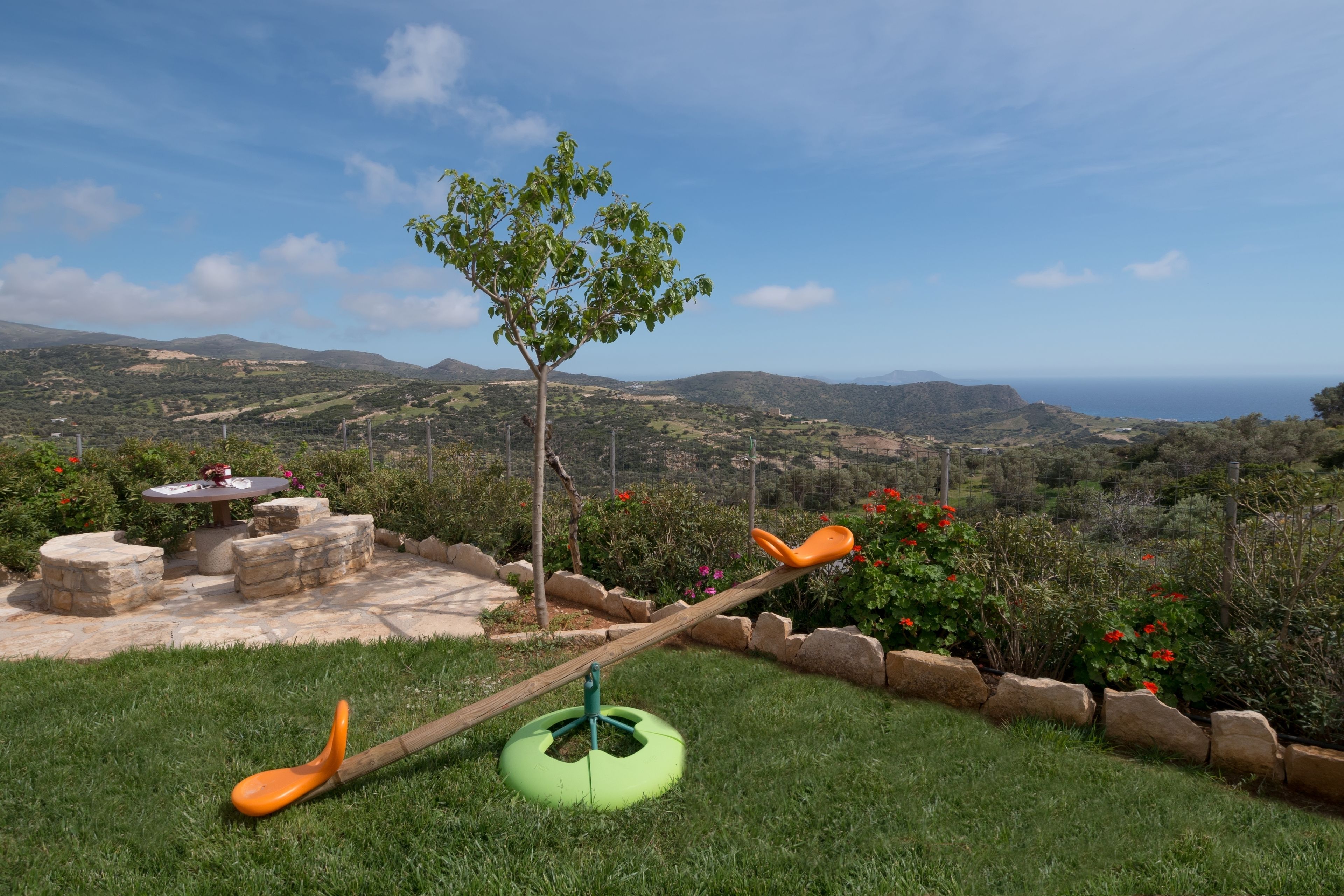 A seesaw with a tree in the middle, a stone table with chairs, and a view of the landscape.