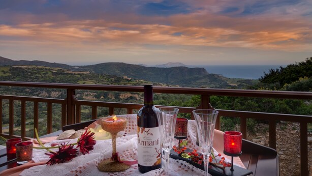 Una mesa dispuesta con una botella de vino, copas y velas en un balcón con vistas a un paisaje montañoso.