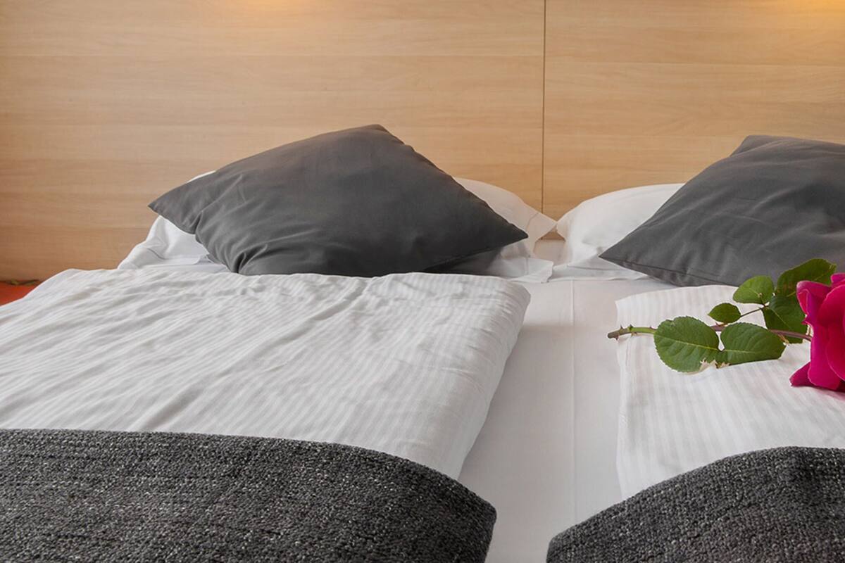A neatly made bed with white and gray bedding, a single rose on the bed, and a wooden headboard with a phone mounted on it.