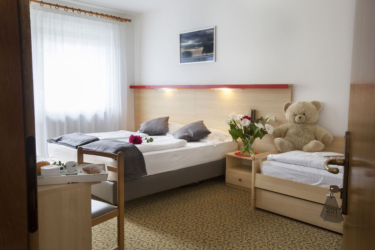 A hotel room with two beds, a wooden headboard, a bedside table with a teddy bear, a tray with breakfast items, and a window with sheer curtains.
