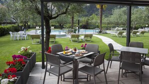 Um pátio externo com vista para a piscina, mobiliado com cadeiras e mesas, rodeado por vegetação e um caminho.