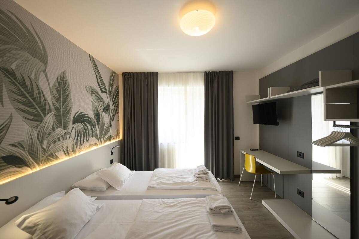 A hotel room with two beds, a desk, a TV, and a large plant mural.