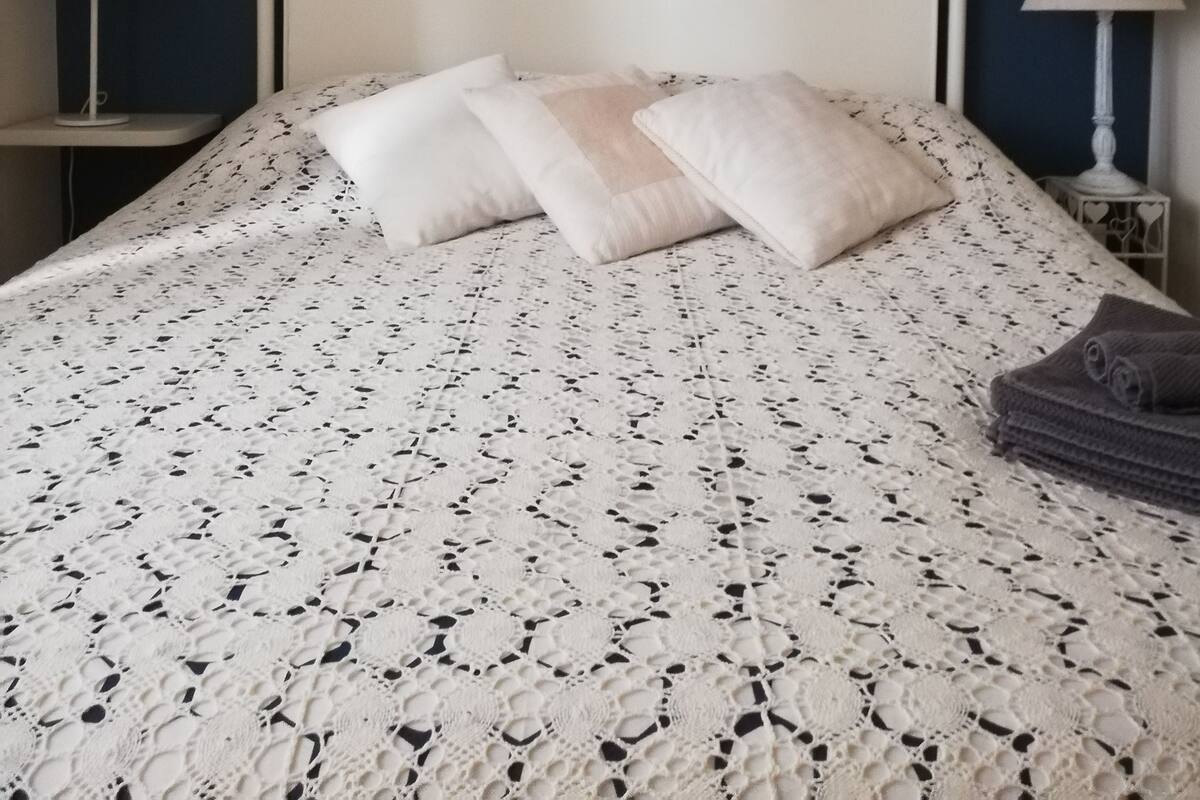 Ein Bett mit weißem Kopfteil und einem gemusterten Überwurf, flankiert von zwei Nachttischen mit Lampen, vor einer dunkelblauen Wand mit zwei gerahmten Spiegeln.