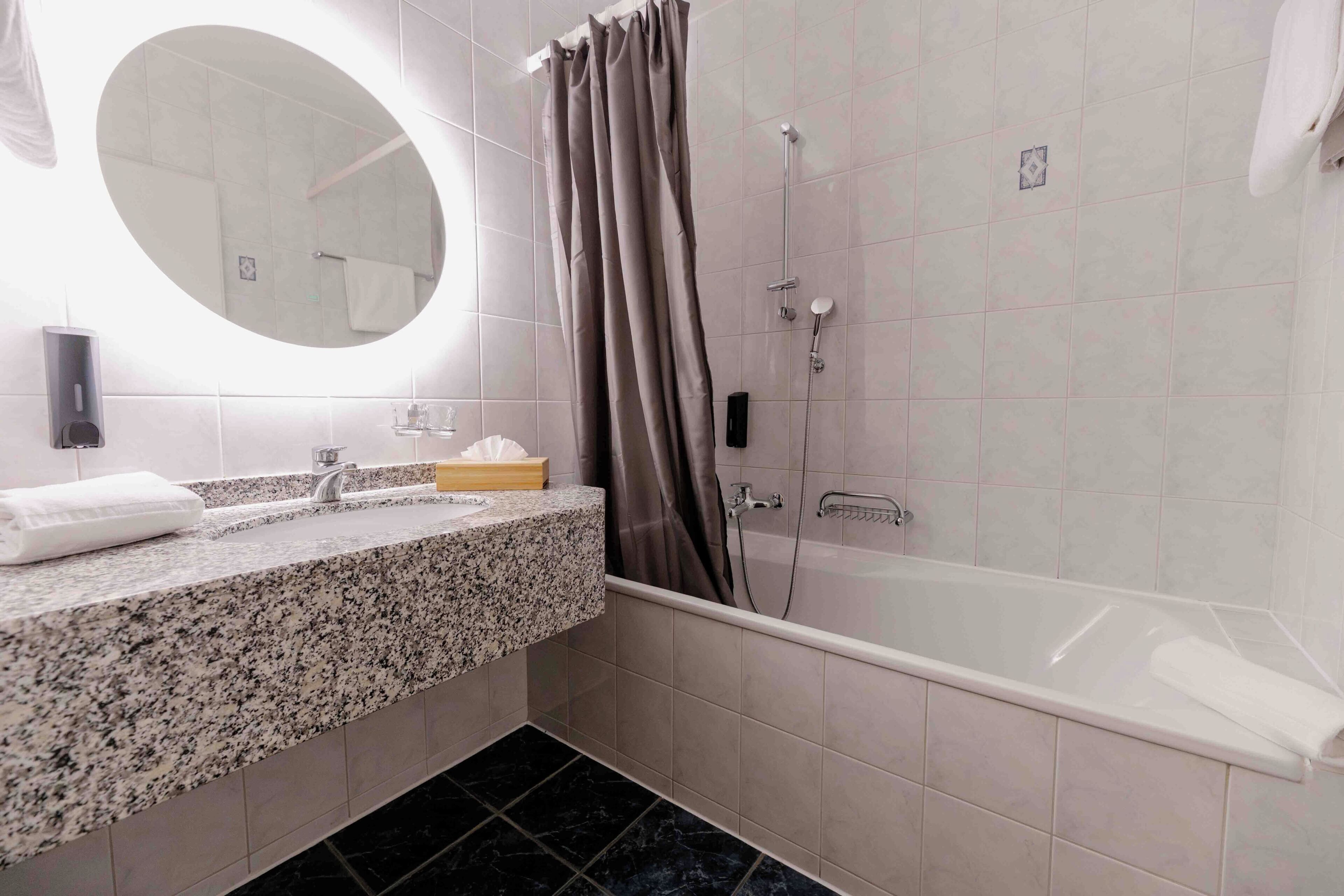 Un baño con encimera de granito, una bañera blanca, ducha con cortina y un espejo redondo.