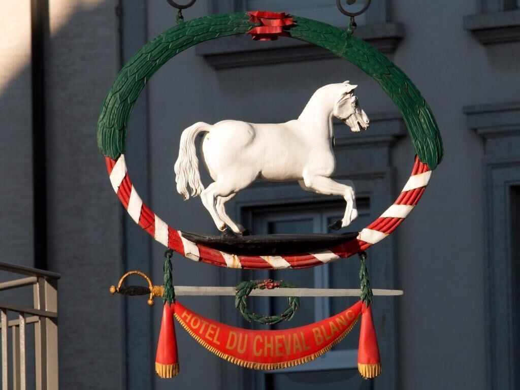 Una estatua de un caballo blanco montada sobre un marco circular con un borde a rayas rojas y blancas, colgando de un arreglo verde. Debajo del caballo, un banderín con la leyenda 'HOTEL DU CHEVAL BLANC'.
