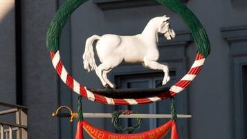 Una estatua de un caballo blanco montada sobre un marco circular con un borde a rayas rojas y blancas, colgando de un arreglo verde. Debajo del caballo, un banderín con la leyenda 'HOTEL DU CHEVAL BLANC'.