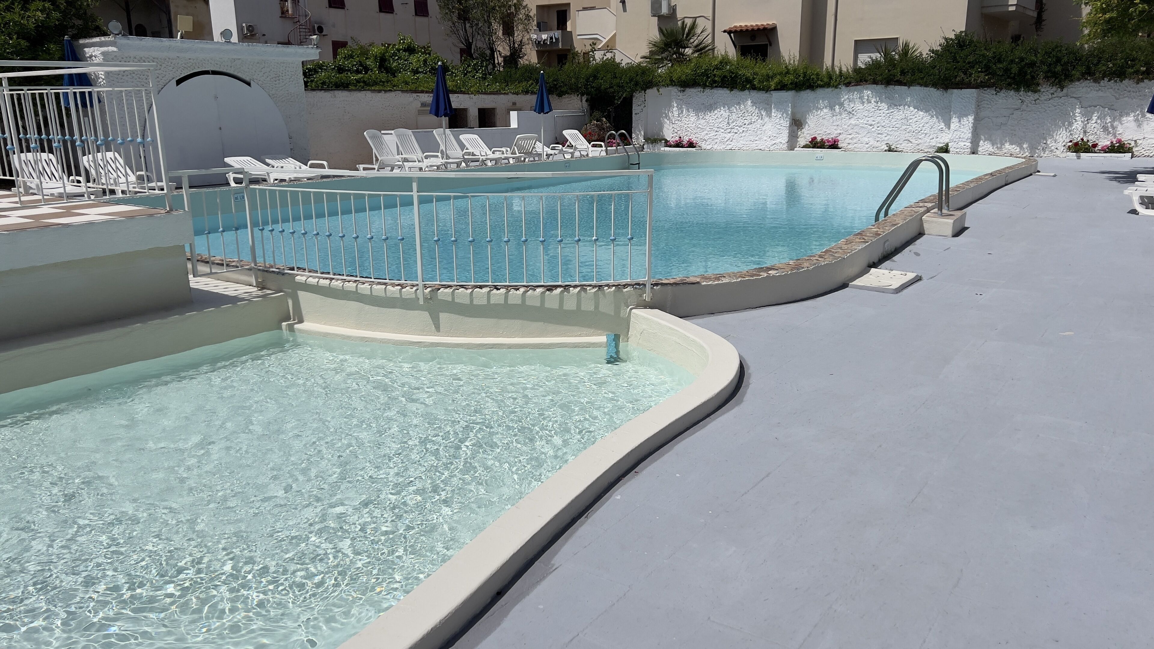 Zona de piscina con agua cristalina, sillas de descanso blancas y una pared blanca con diseño texturizado.