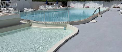 Zona de piscina con agua cristalina, sillas de descanso blancas y una pared blanca con diseño texturizado.