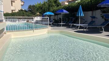 Una piscina con agua cristalina, rodeada de sillas de descanso y sombrillas, y una valla blanca.