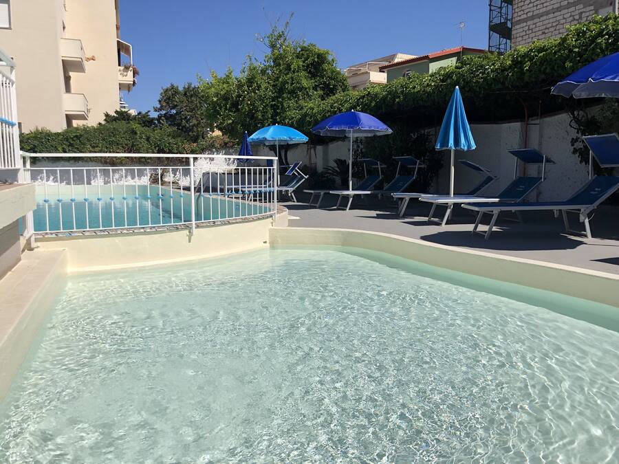 Una piscina con agua cristalina, rodeada de sillas de descanso y sombrillas, y una valla blanca.