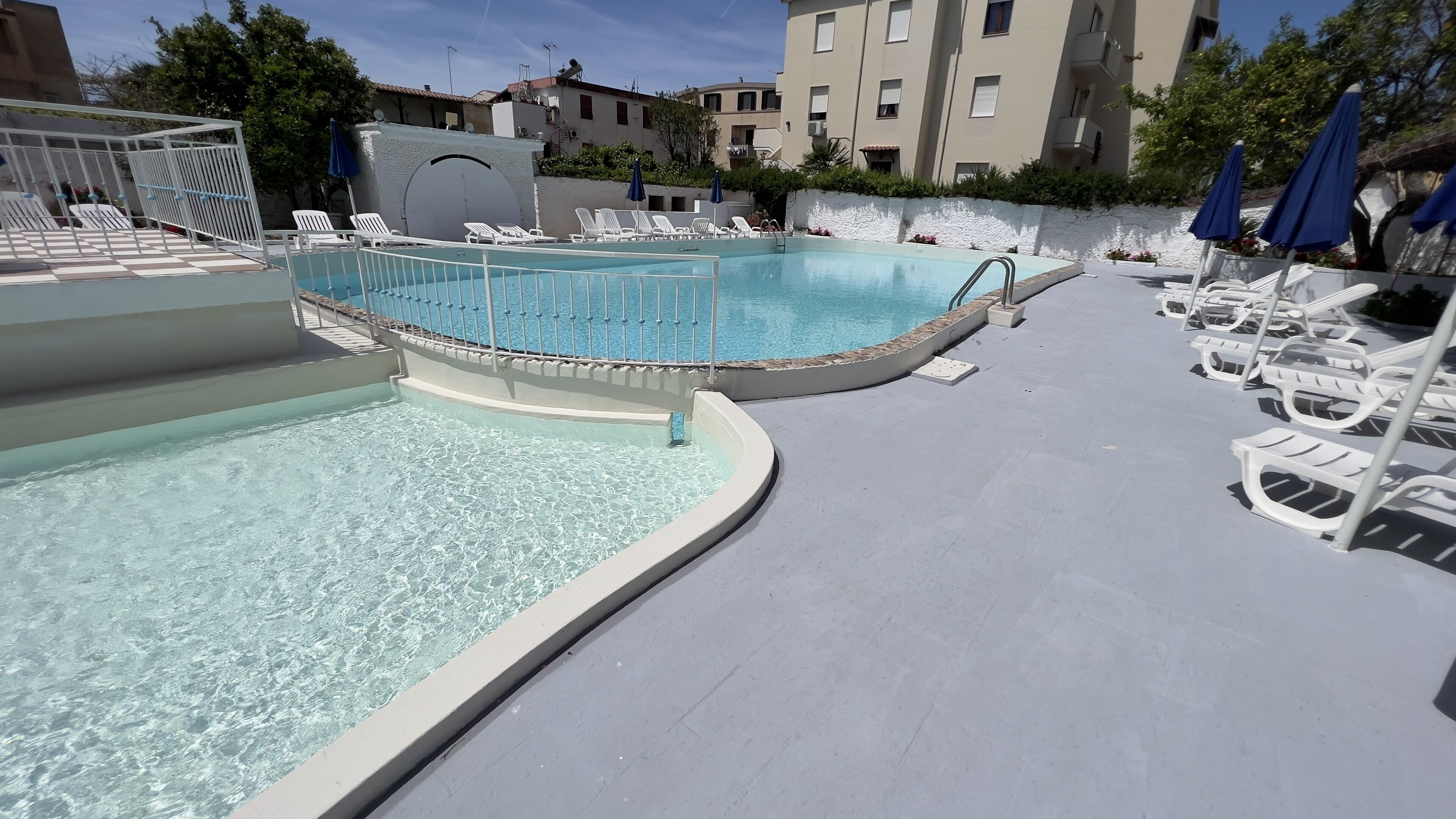 A swimming pool with a curved edge, surrounded by white lounge chairs and blue umbrellas.