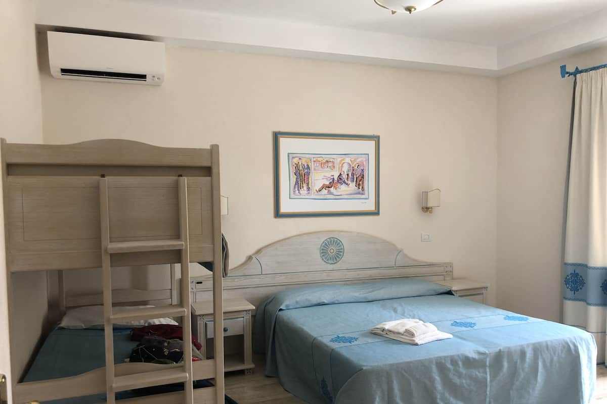 A bedroom with a bunk bed, a desk, a bed with a blue bedspread, a wall-mounted air conditioner, a framed artwork, and a window with curtains.