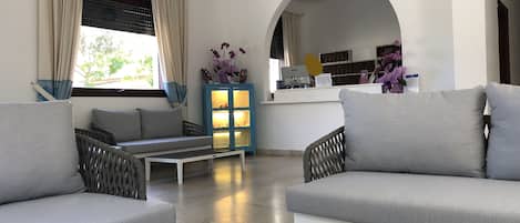 A modern living room with grey sofas, a coffee table with brochures, a blue cabinet with glass doors, and a reception desk with a computer and flowers.