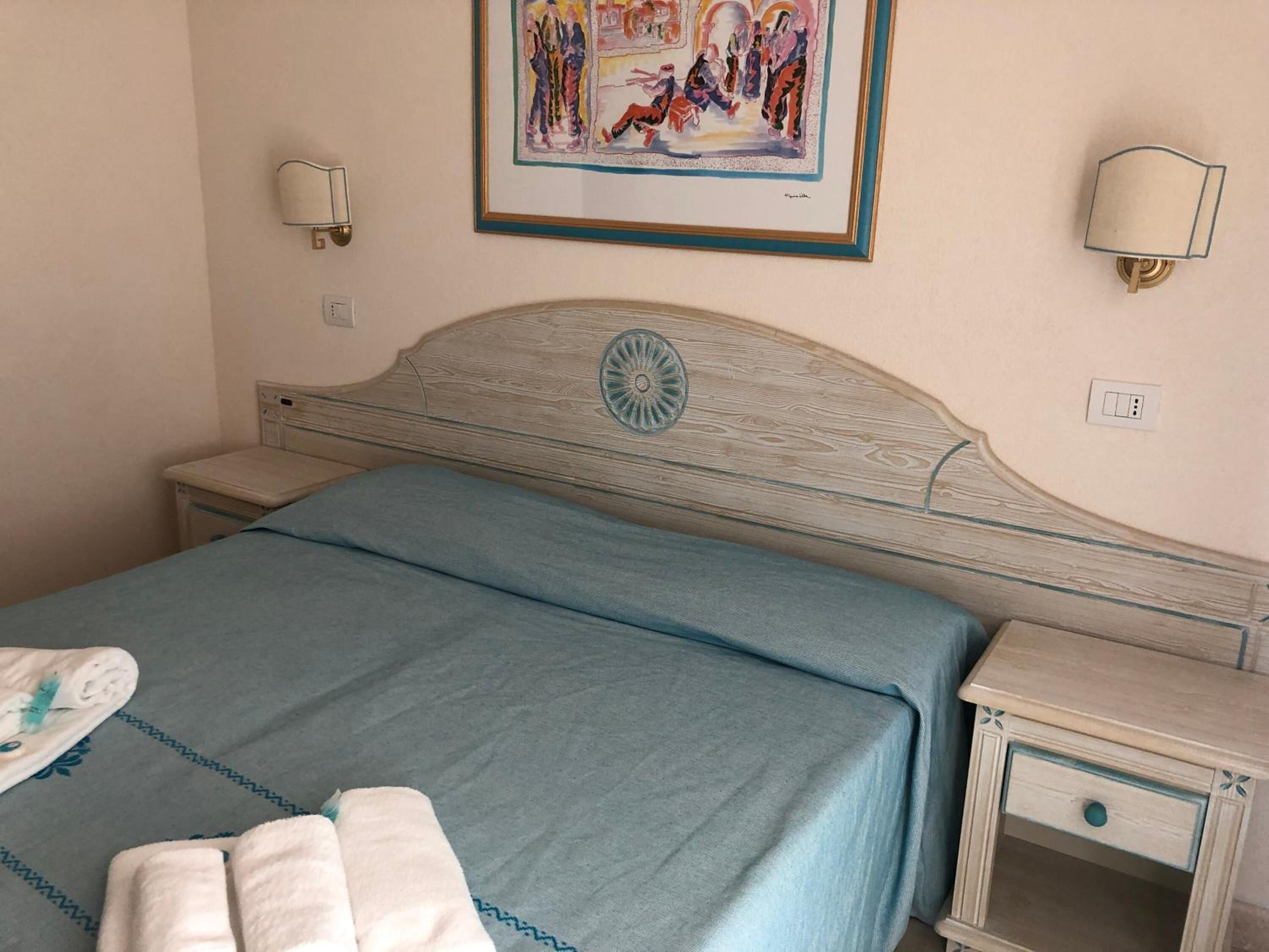A bedroom with a bed, a nightstand, a wall-mounted lamp, and a painting.