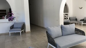 Un espacio moderno para relajarse con un sofá gris, una silla de mimbre y un sillón blanco. Hay una pequeña mesa con un libro y un jarrón de flores. Las paredes son blancas y el piso está cubierto de baldosas.