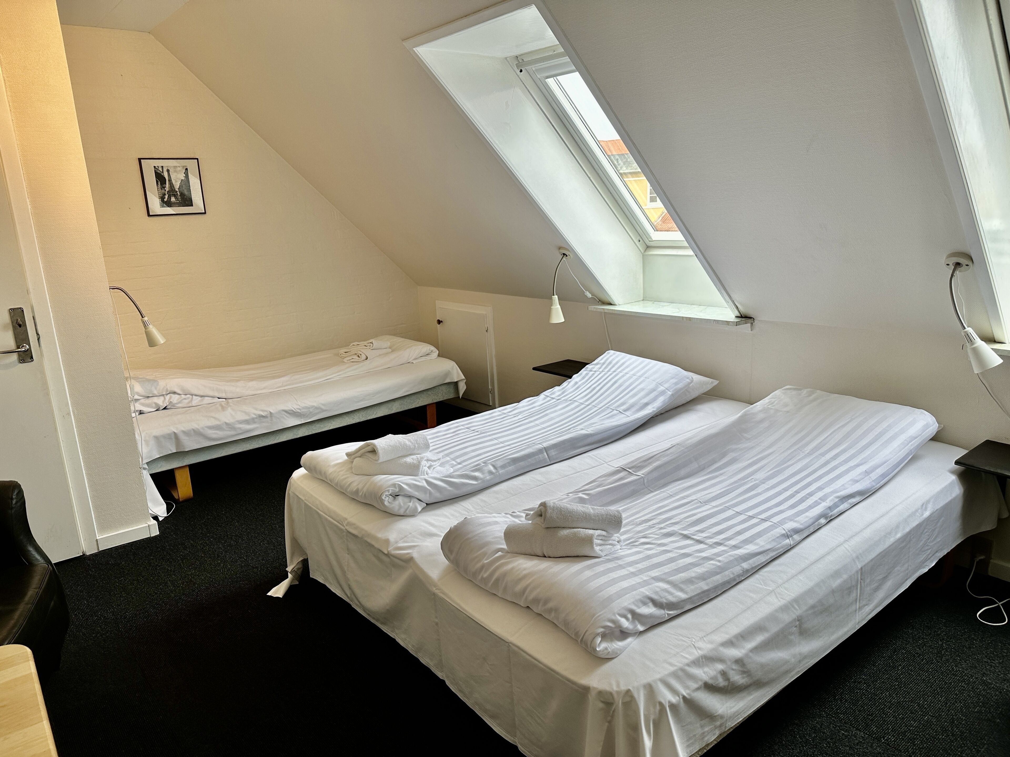 Une chambre de dortoir avec deux lits simples, une lucarne, un petit bureau et un tableau accroché au mur.