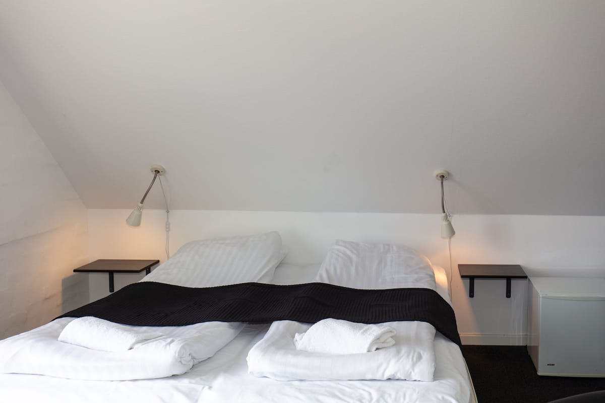 Una cama con dos camas individuales con ropa de cama blanca y una manta negra, dos mesitas de noche con lámparas y un techo blanco.