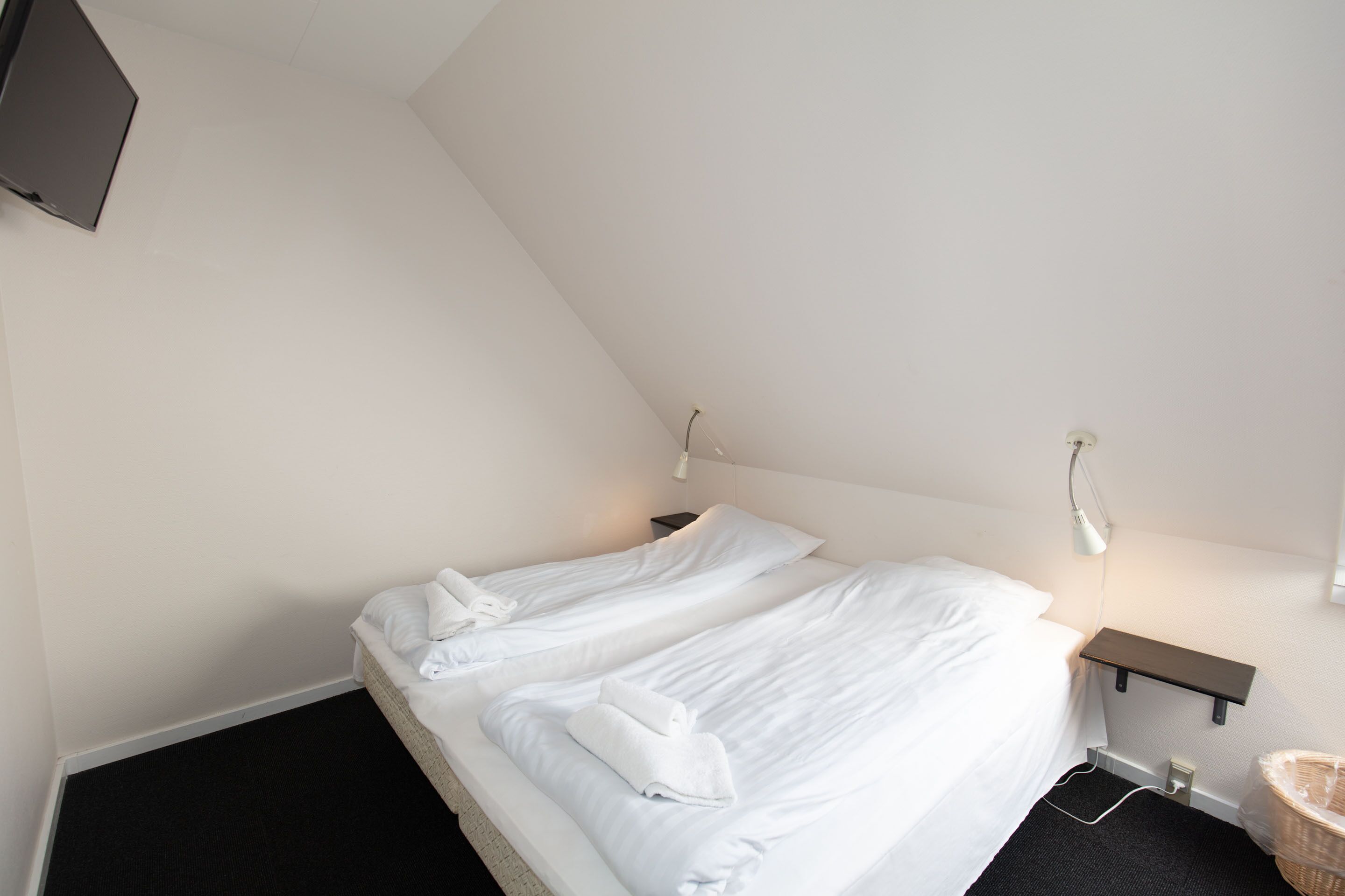 Una habitación de hotel con dos camas, una mesita de noche, un televisor montado en la pared y un estante pequeño.