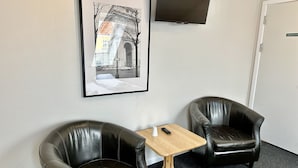 Une pièce comprenant deux fauteuils en cuir noir, une petite table en bois avec une télécommande, et un tableau représentant une scène de rue accroché au mur.