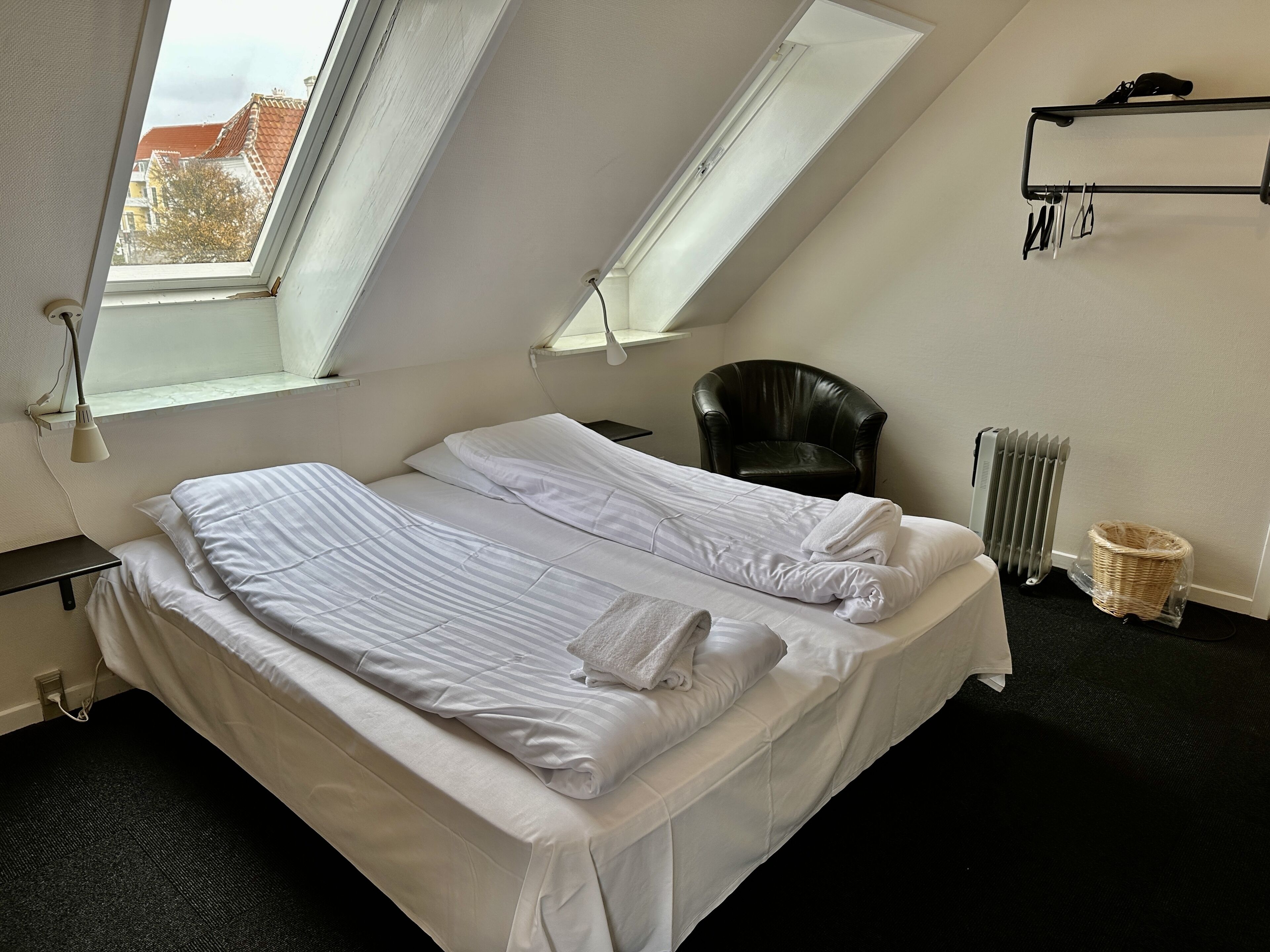 A neatly made bed in a room with a skylight, a black chair, a radiator, and a wall-mounted rack with items.