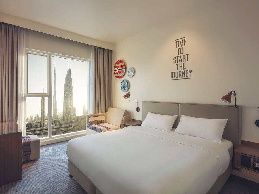 A hotel room with a large bed, a desk, a chair, a view of a city skyline, and a motivational wall quote.