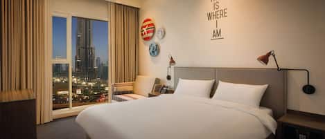 A hotel room with a large bed, a desk, a chair, a nightstand, a lamp, a wall clock, and a view of the city at night.