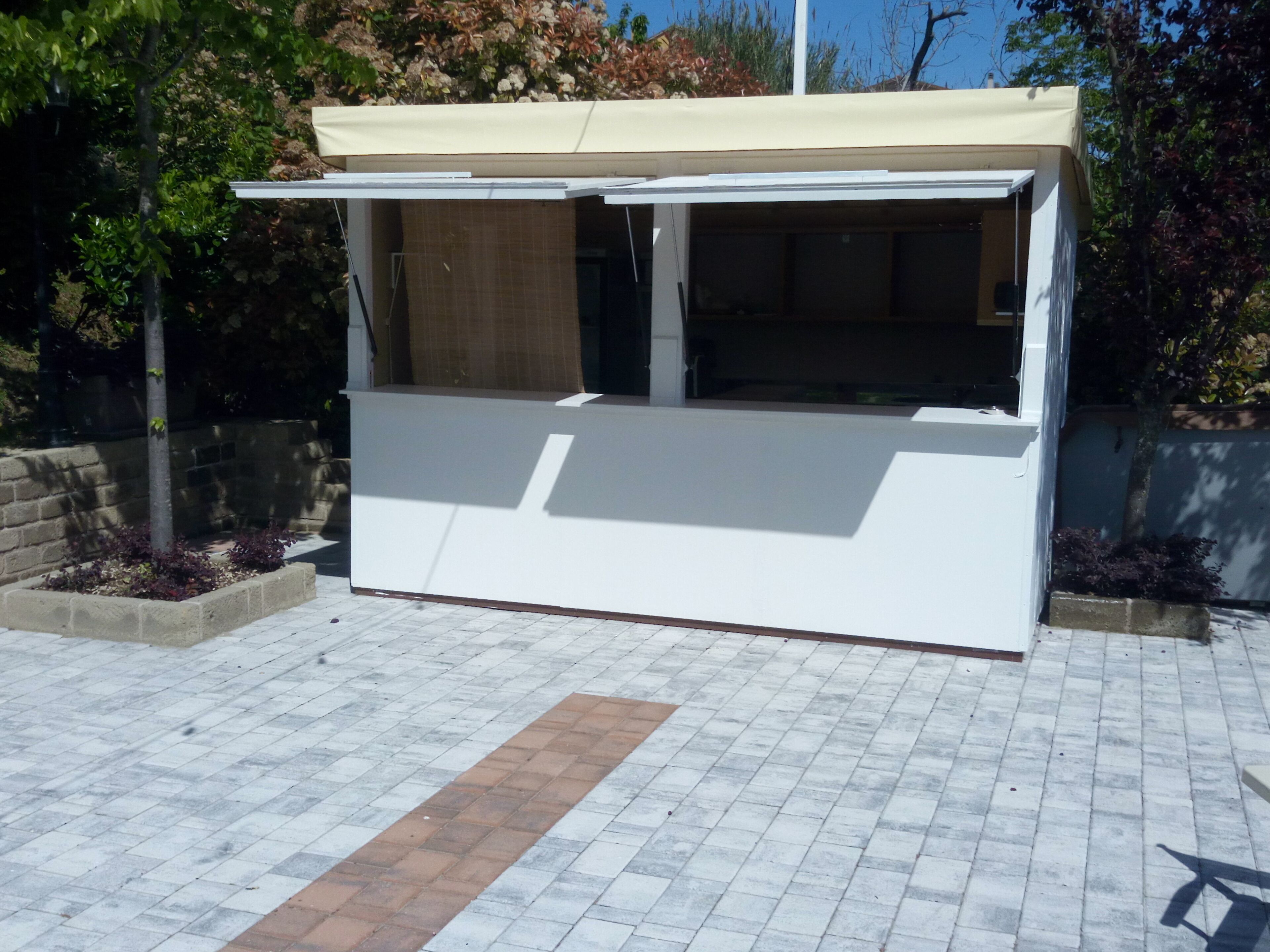 A white outdoor kiosk with a beige awning, surrounded by a paved area with brick and stone tiles, and adjacent to a low stone wall with plants.