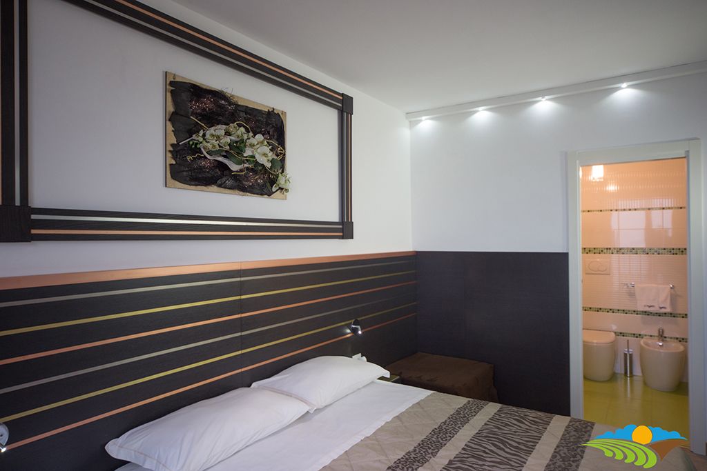 Un dormitorio con cama, cabecera a rayas horizontales, una obra de arte colgada en la pared y un baño visible a través de una puerta abierta.