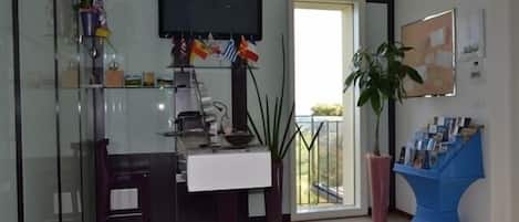 A modern living room with a glass partition, a flat-screen TV, a desk with a computer, a chair, a potted plant, and a blue magazine rack.