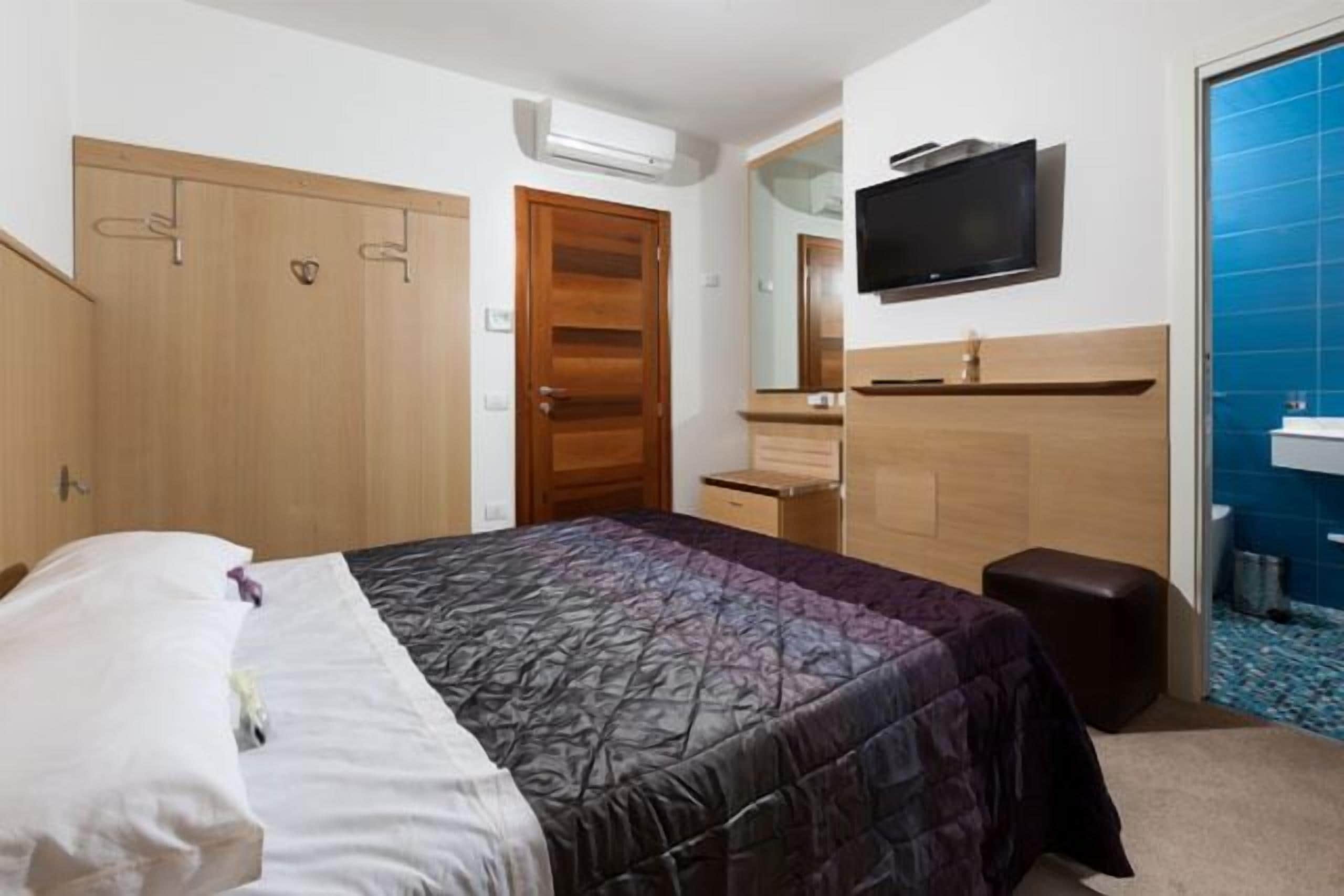 A bedroom with a bed, a television, a mirror, a wardrobe, and a bathroom with blue tiles.