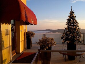 Een balkon met uitzicht op een waterpartij en bergen, een kerstboom en een rode luifel.