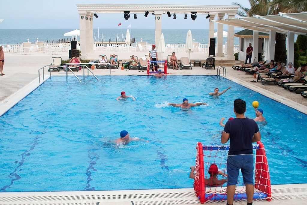 Ein Schwimmbecken mit einem laufenden Wasserballspiel, umgeben von Liegestühlen und Sonnenschirmen.