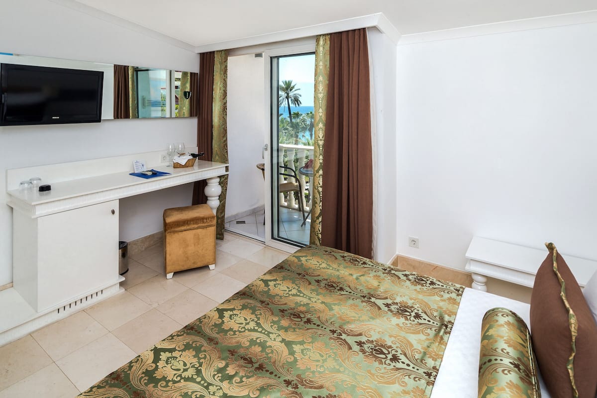 A hotel room with a bed, a desk, a television, and a balcony with a view of palm trees.